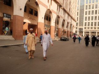 People walking on a bustling street in Makkah, showcasing traditional attire and Middle Eastern architecture.